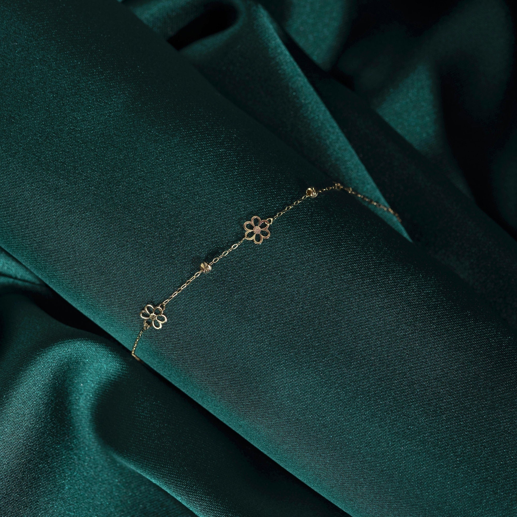 Fine gold-colored chain bracelet with two open flower motifs and small bead accents, draped diagonally over dark green fabric.