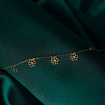 Fine gold-tone chain bracelet with four dangling open-petal flower charms, laid diagonally across dark teal fabric.