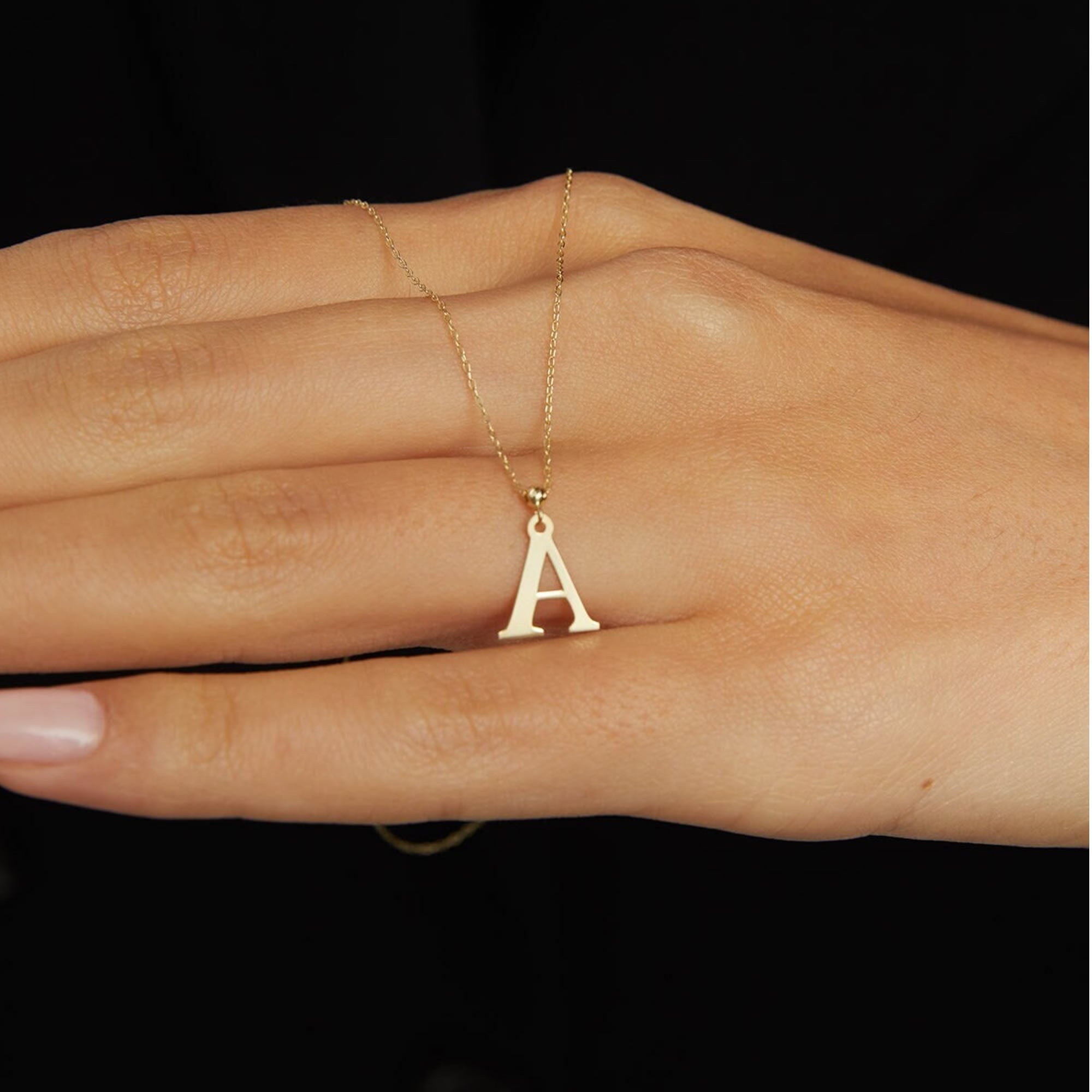Gold-colored necklace with a fine chain and a capital A pendant resting across a hand against a black background.