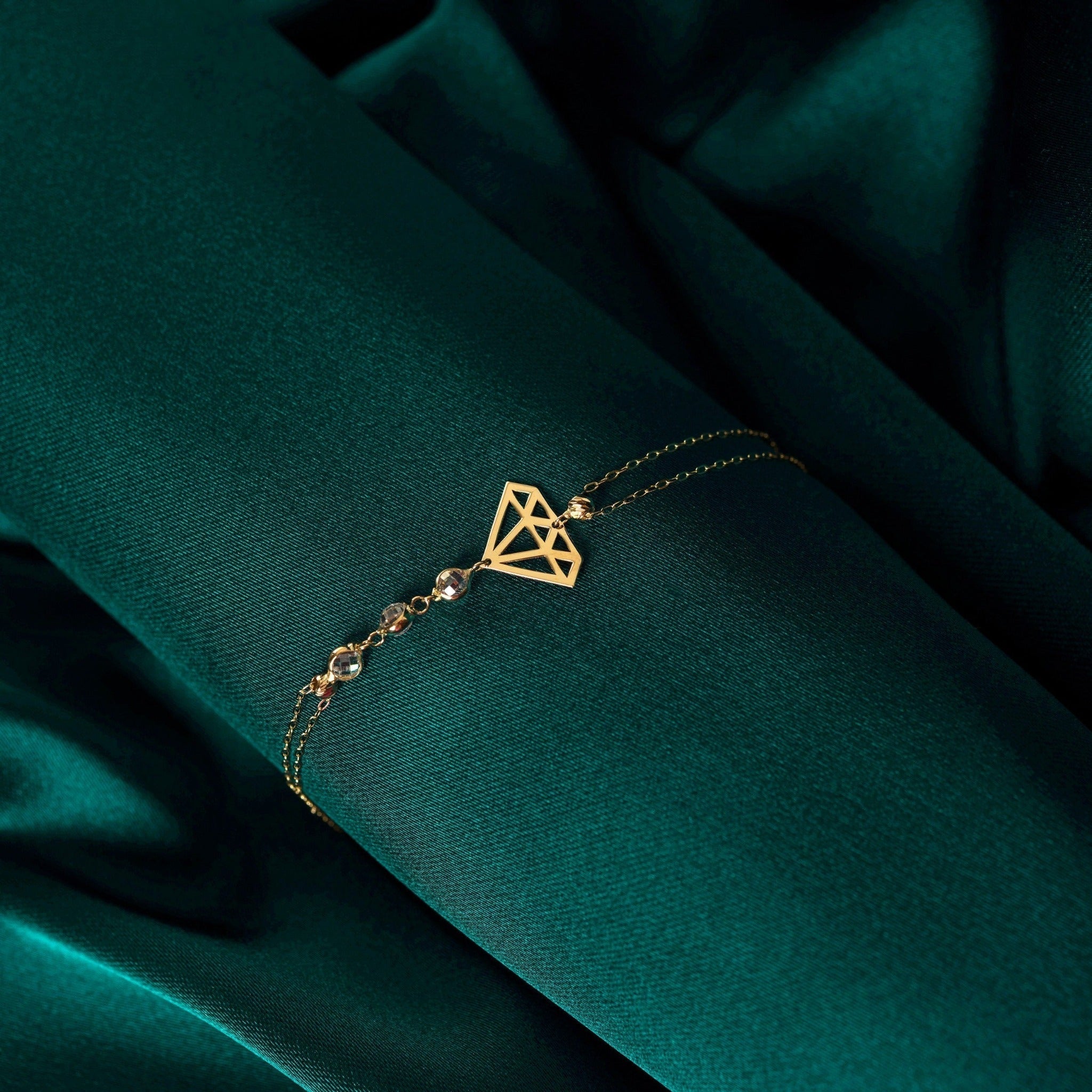 Gold-tone double-chain bracelet with an openwork diamond-shaped motif and faceted beads on dark teal fabric.
