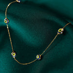 Fine gold-colored chain necklace with spaced blue, white and yellow eye motifs, draped over dark green fabric.