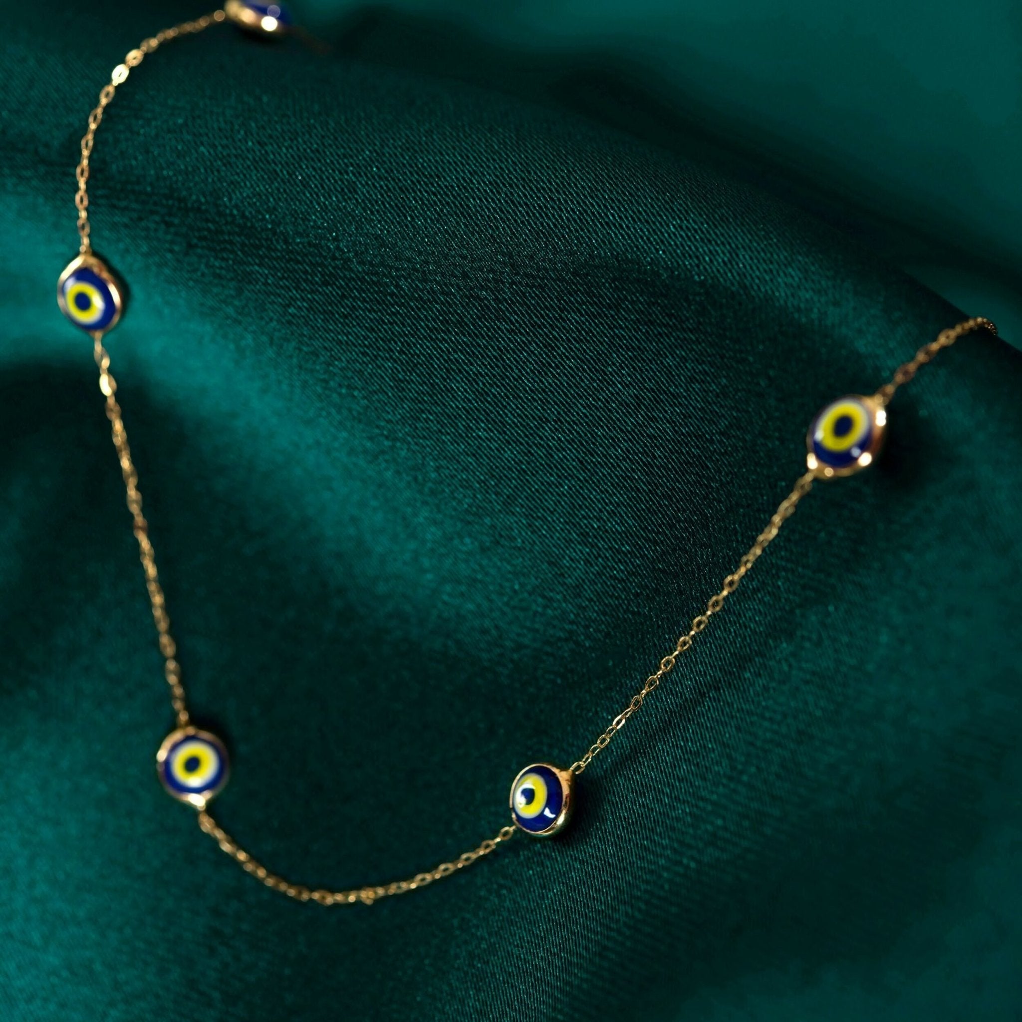 Fine gold-colored chain necklace with spaced blue, white and yellow eye motifs, draped over dark green fabric.