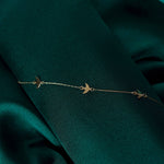 Fine gold-toned chain bracelet with three flat bird charms, laid diagonally across dark green folded fabric.
