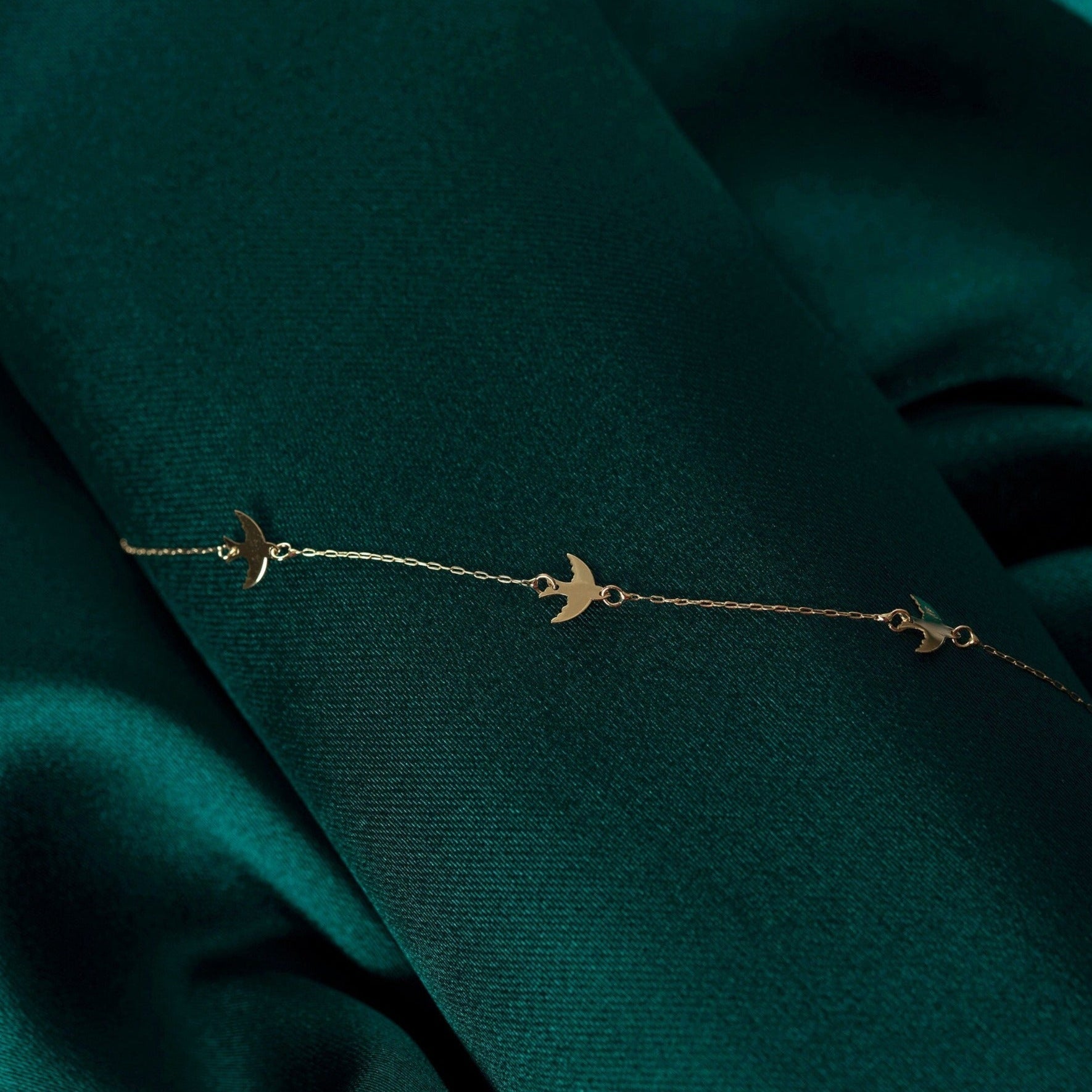 Fine gold-toned chain bracelet with three flat bird charms, laid diagonally across dark green folded fabric.