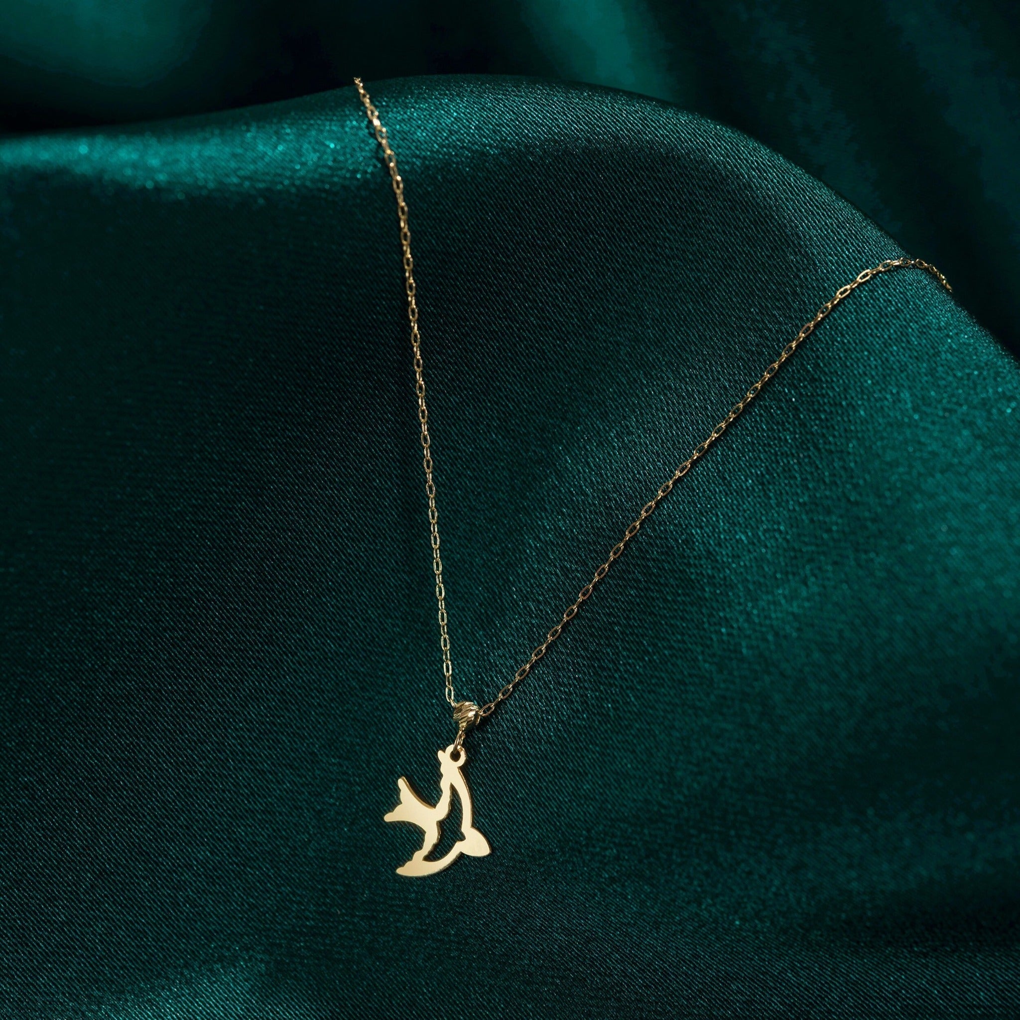 Gold-toned necklace with a fine oval-link chain and a cutout flying bird pendant resting on dark teal fabric.