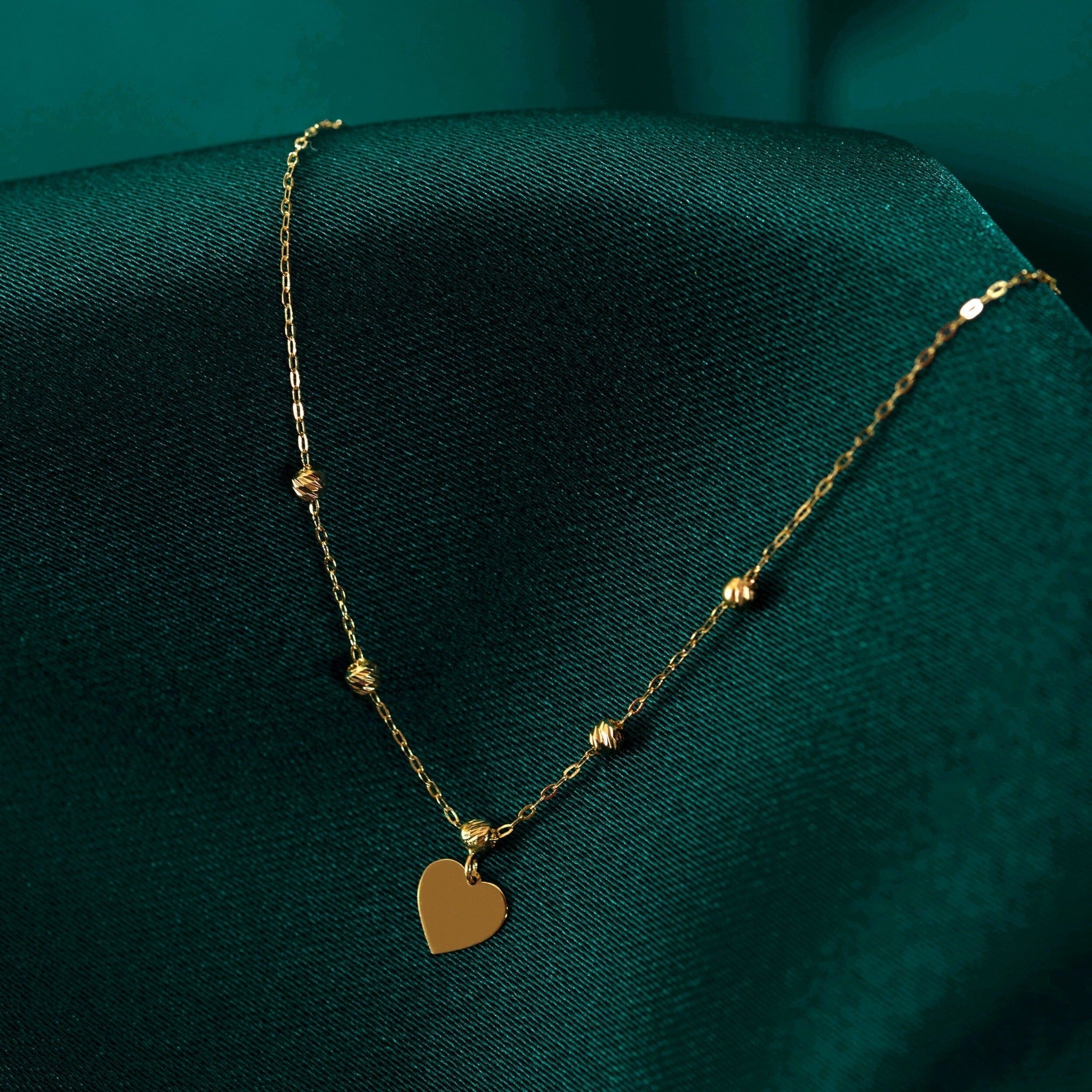 Gold-colored necklace with a flat heart pendant and textured round beads along a fine link chain, draped over dark green fabric.
