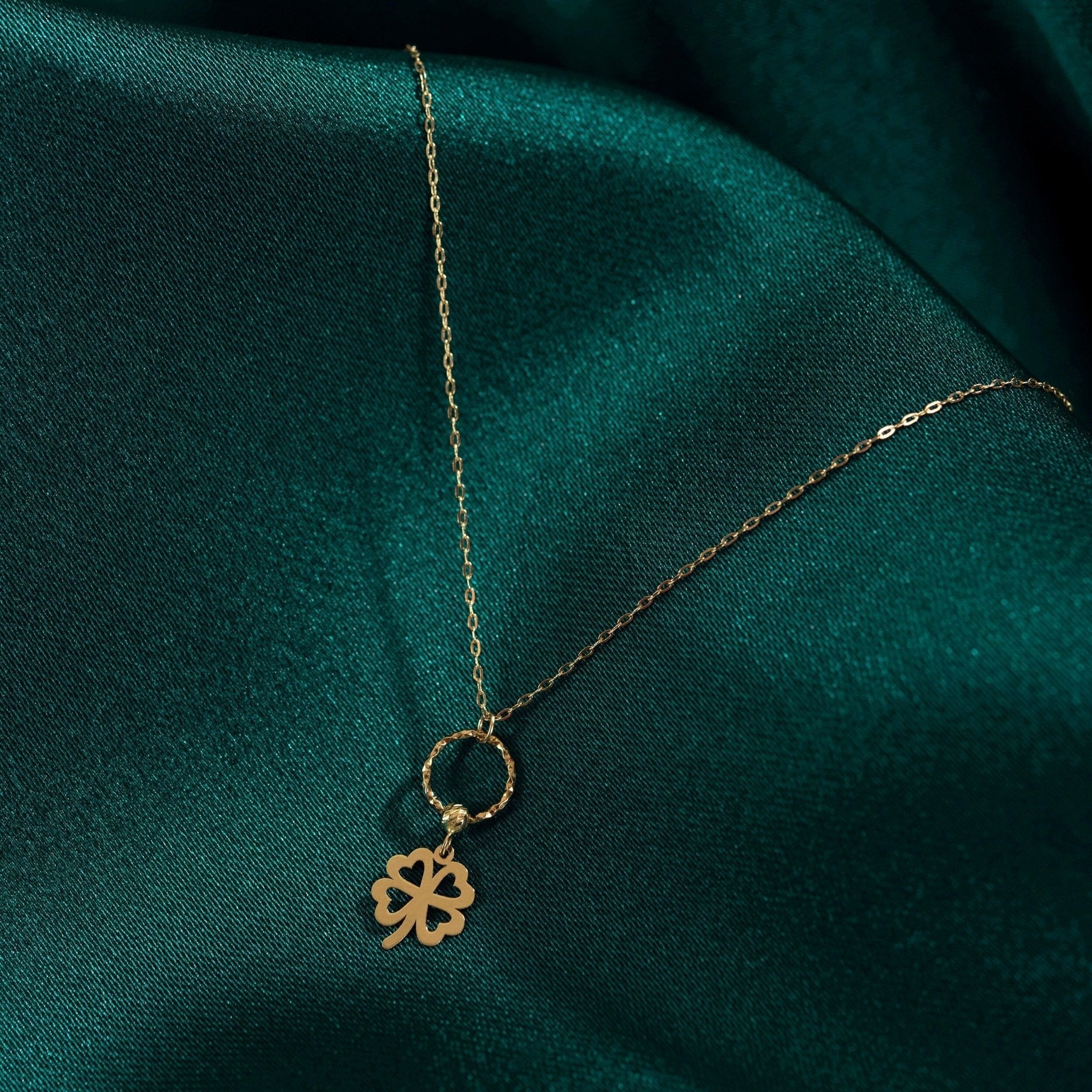 Gold-tone chain necklace with an openwork four-leaf clover hanging from a textured ring, resting on dark green fabric.