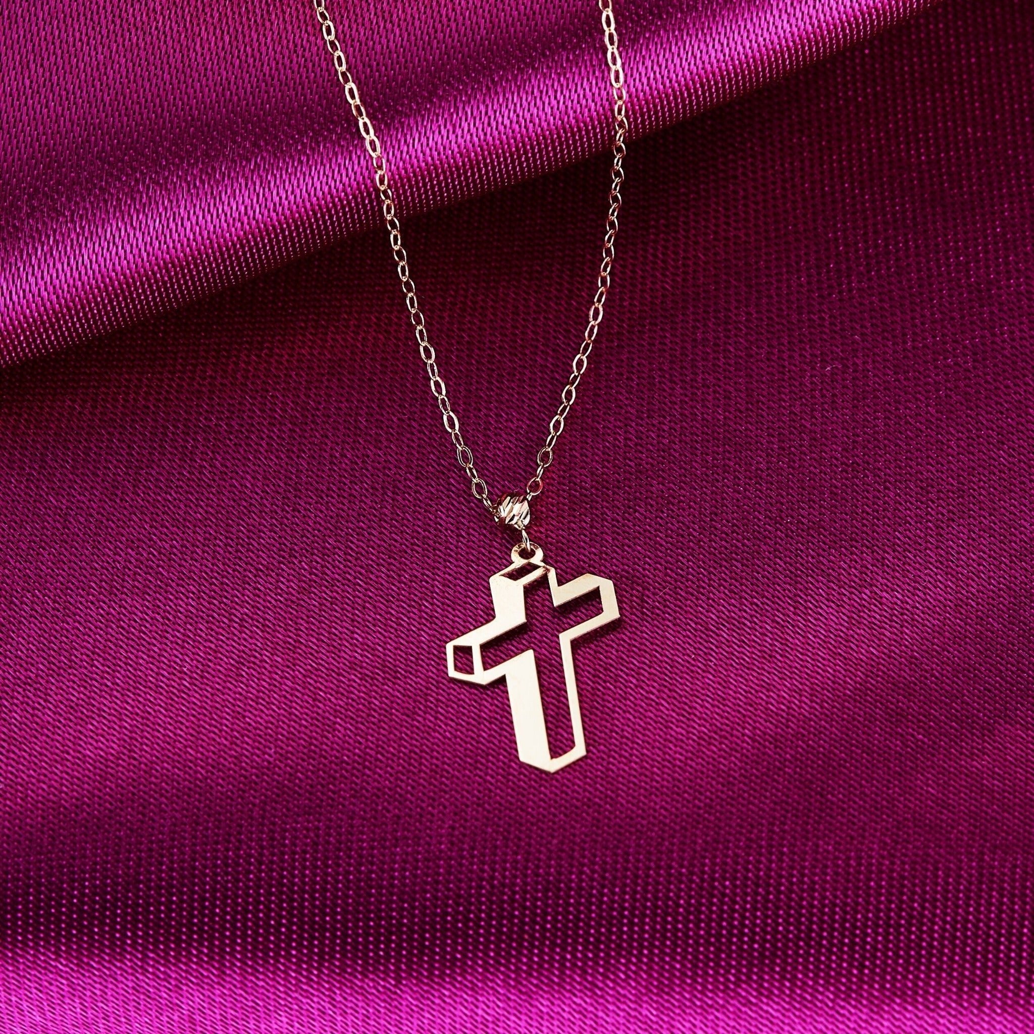 Gold-tone necklace with an open geometric cross pendant and fine oval-link chain on folded magenta fabric.