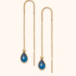 Pair of gold-tone threader earrings with fine chains, curved tops and faceted blue teardrop pendants on a cream background.