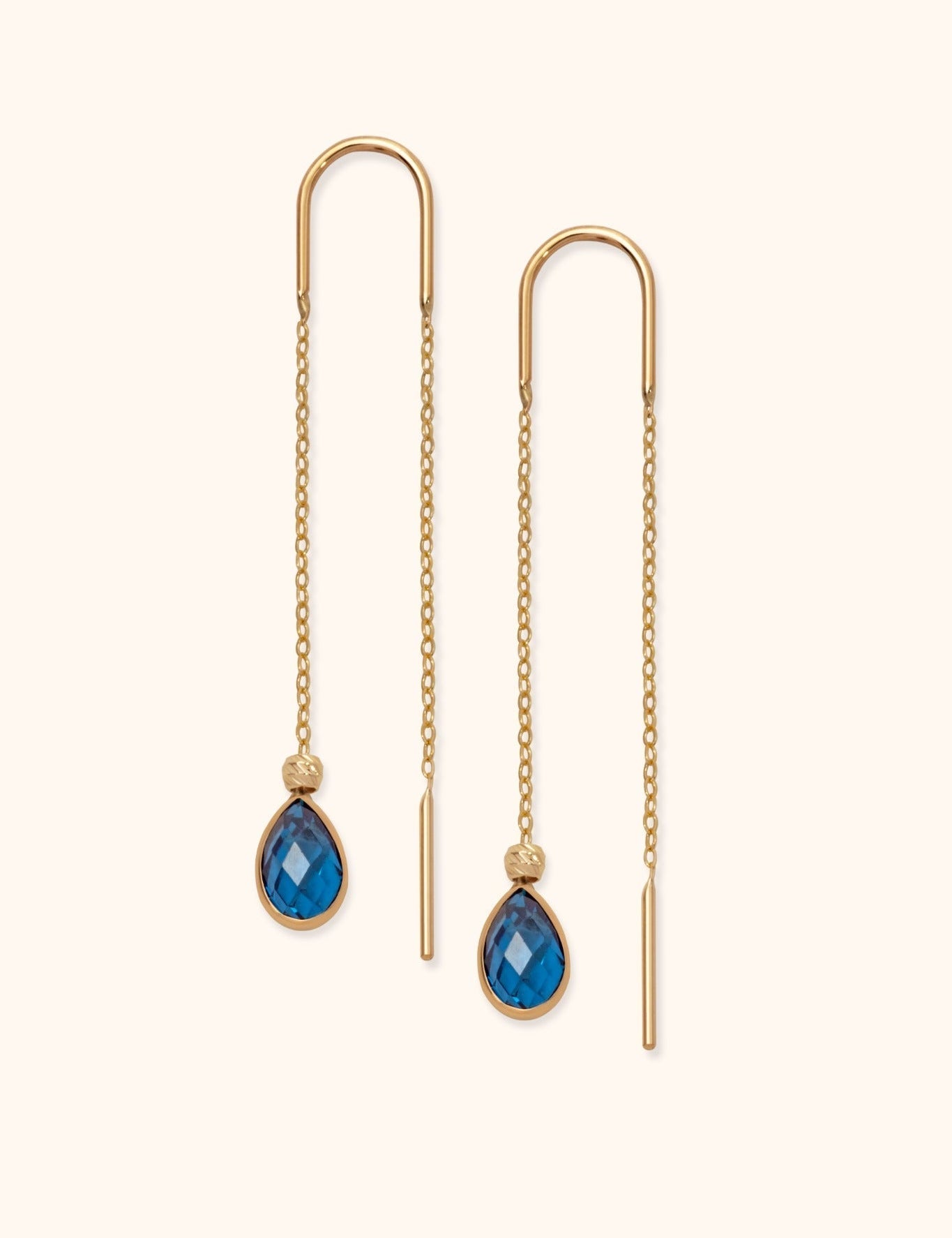 Pair of gold-tone threader earrings with fine chains, curved tops and faceted blue teardrop pendants on a cream background.