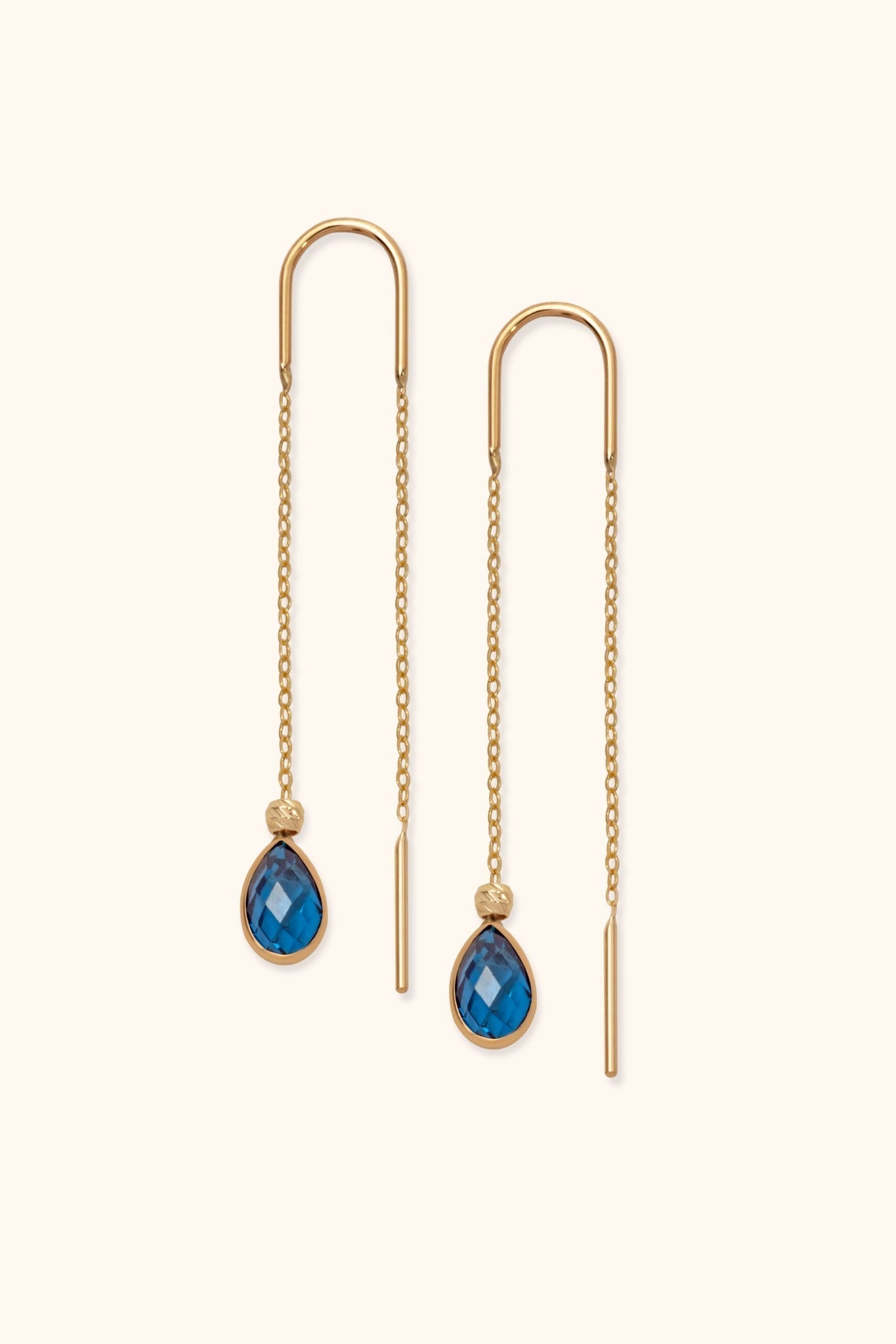 Pair of gold-tone threader earrings with fine chains, curved tops and faceted blue teardrop pendants on a cream background.