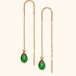 Pair of gold-tone threader earrings with fine chains, curved tops and faceted green teardrop pendants on a cream background.