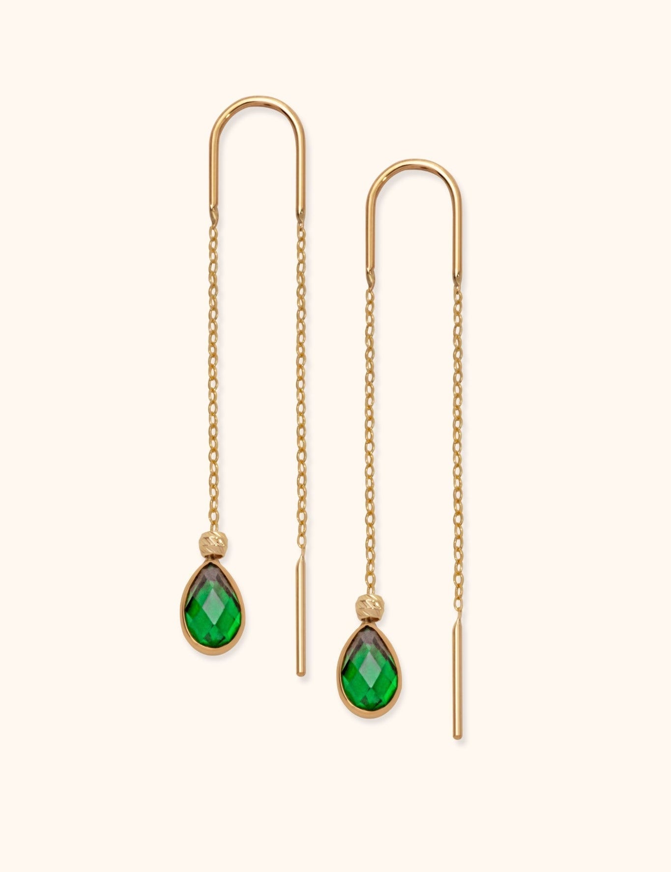 Pair of gold-tone threader earrings with fine chains, curved tops and faceted green teardrop pendants on a cream background.
