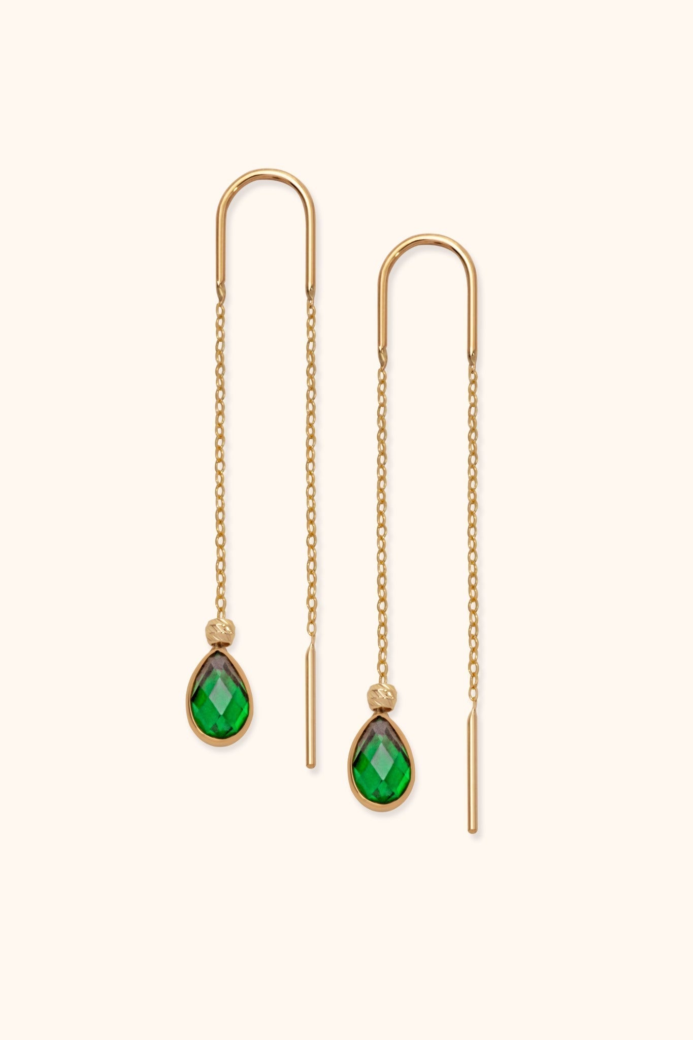 Pair of gold-tone threader earrings with fine chains, curved tops and faceted green teardrop pendants on a cream background.