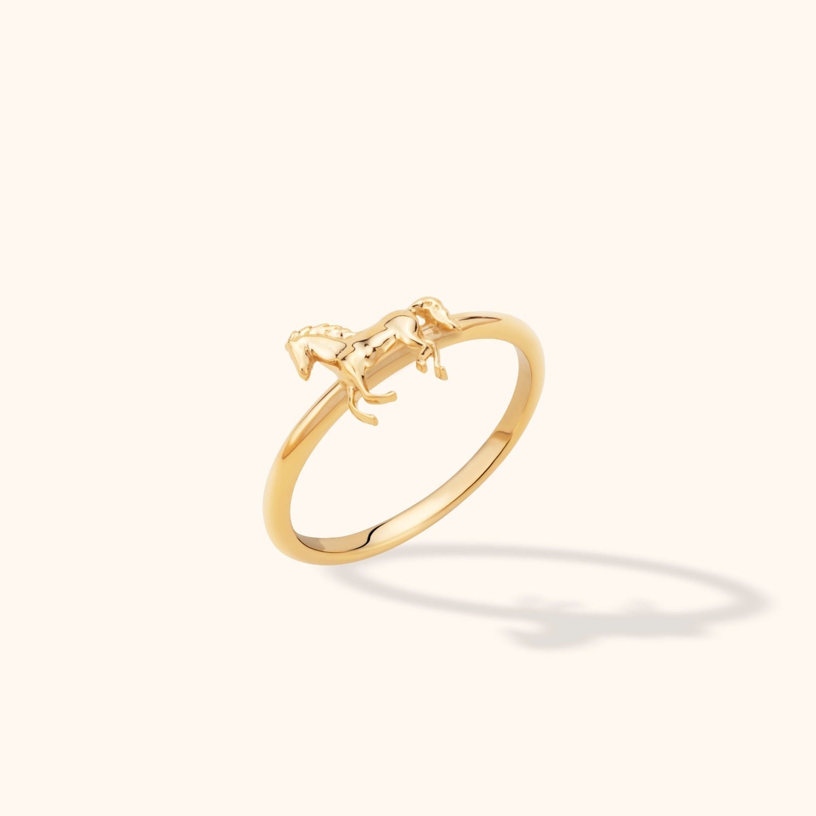Polished gold-toned ring with a slim band and a raised full-body horse figure, shown at an angle on a cream background.