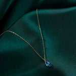 Gold-toned oval-link necklace with a faceted blue teardrop pendant resting on folded dark teal fabric.