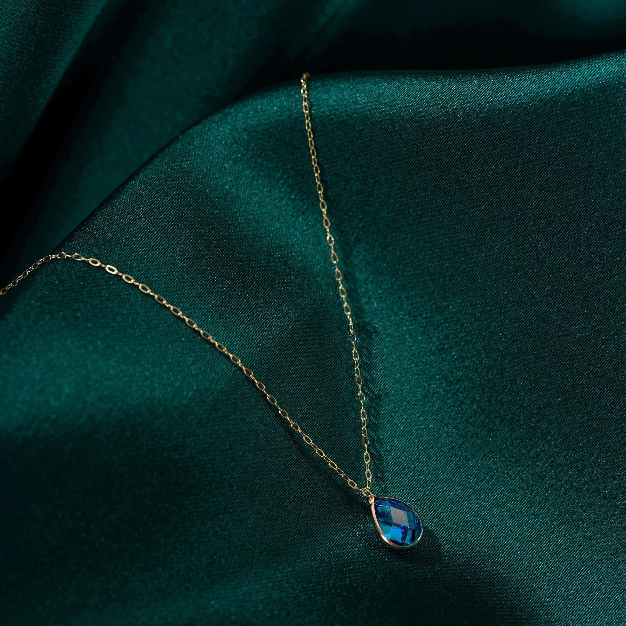Gold-toned oval-link necklace with a faceted blue teardrop pendant resting on folded dark teal fabric.