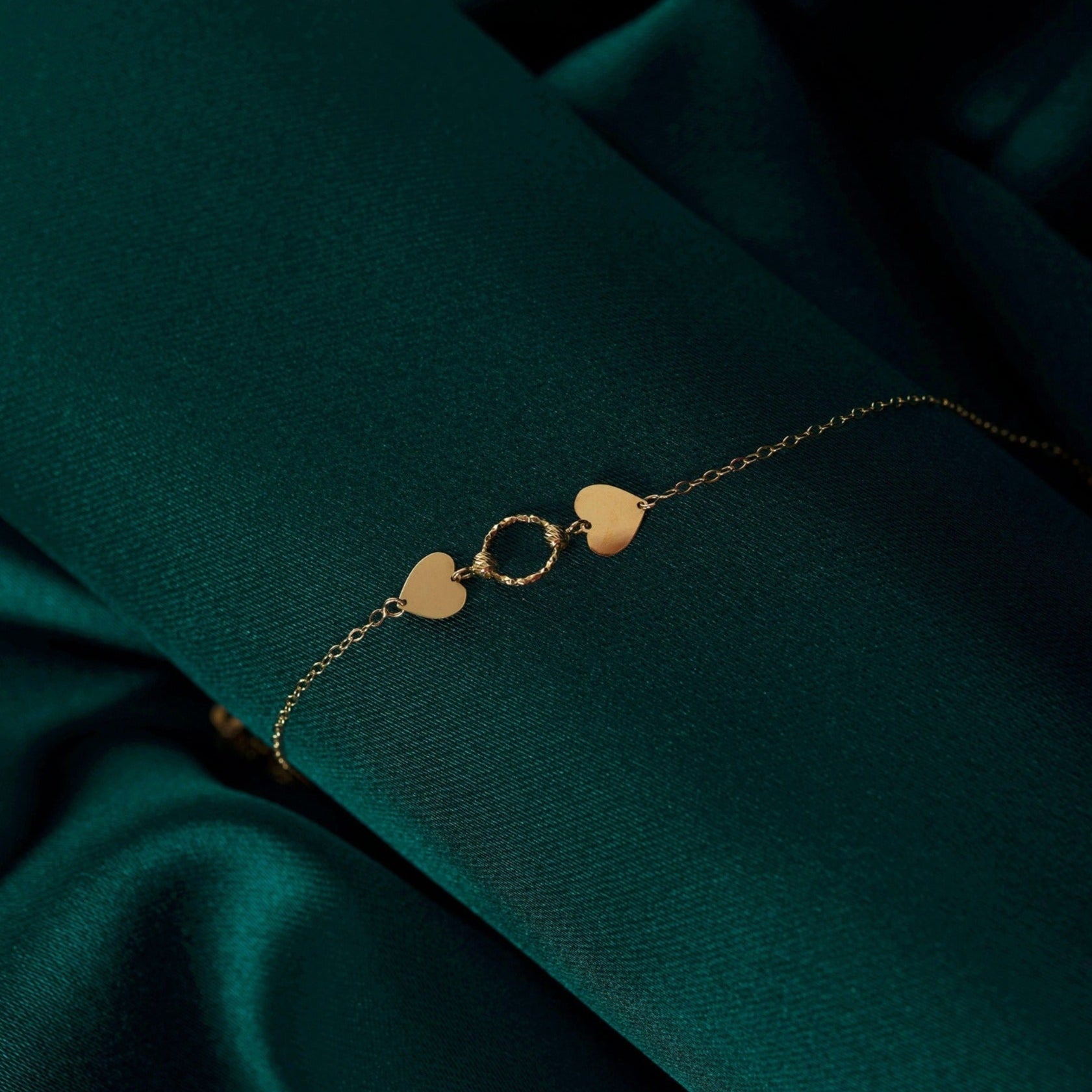 Gold-colored chain bracelet with two flat hearts flanking a twisted circular link, displayed on dark green fabric.