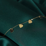 Gold-toned fine chain bracelet with flat, dangling heart charms, laid diagonally across dark green fabric.