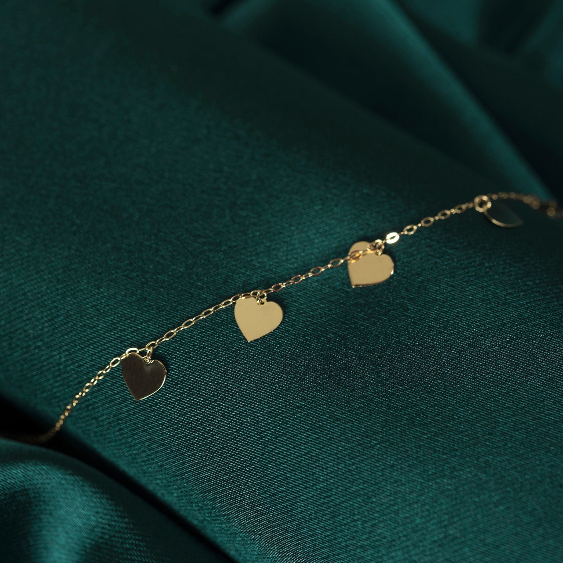 Gold-toned fine chain bracelet with flat, dangling heart charms, laid diagonally across dark green fabric.