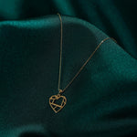 Gold-toned necklace with a geometric cutout heart pendant and fine oval-link chain resting on dark teal fabric.