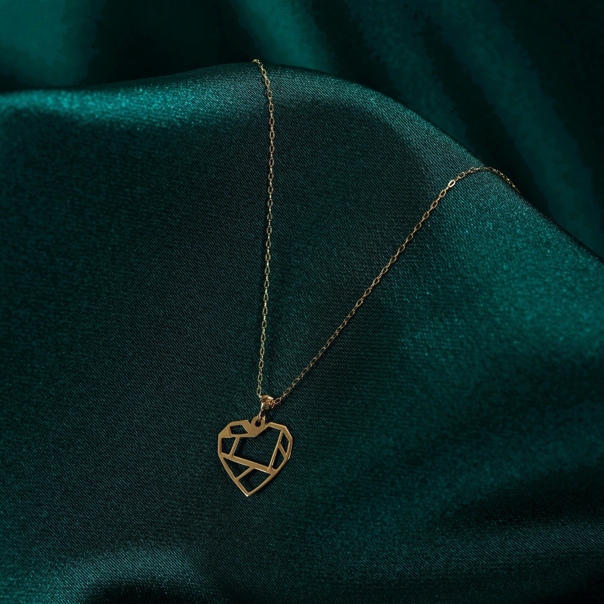 Gold-toned necklace with a geometric cutout heart pendant and fine oval-link chain resting on dark teal fabric.