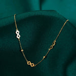 Fine gold-toned chain necklace with three infinity-shaped links and two small beads, draped over dark teal fabric.
