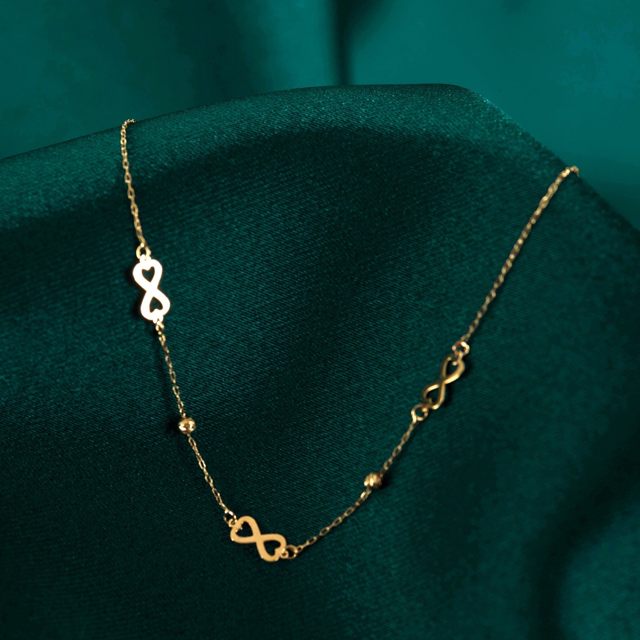 Fine gold-toned chain necklace with three infinity-shaped links and two small beads, draped over dark teal fabric.