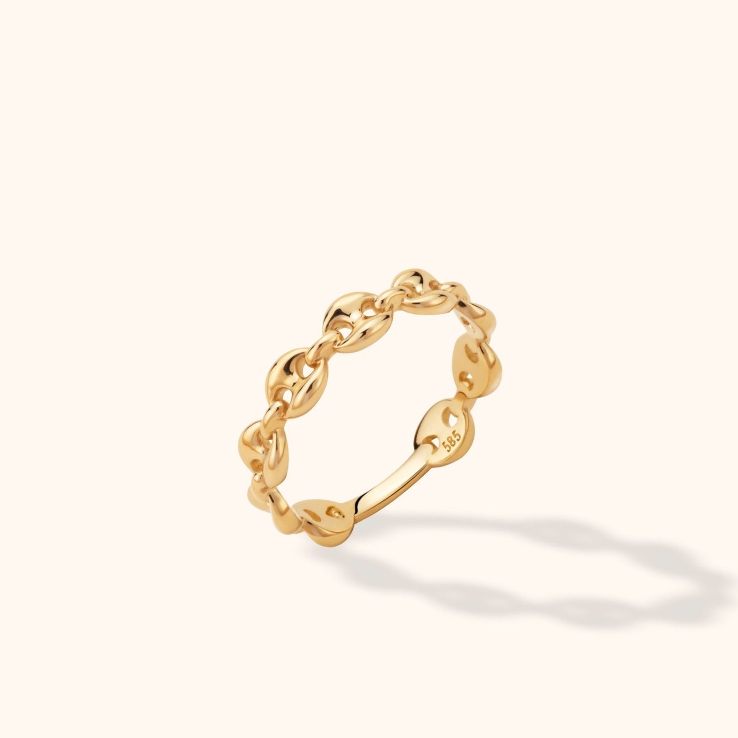 Polished gold-colored ring with interlocking oval chain links, shown tilted on a cream background with a soft shadow.