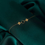 Fine gold-colored chain bracelet with two clover-shaped details and a textured circle, laid diagonally on dark green fabric.