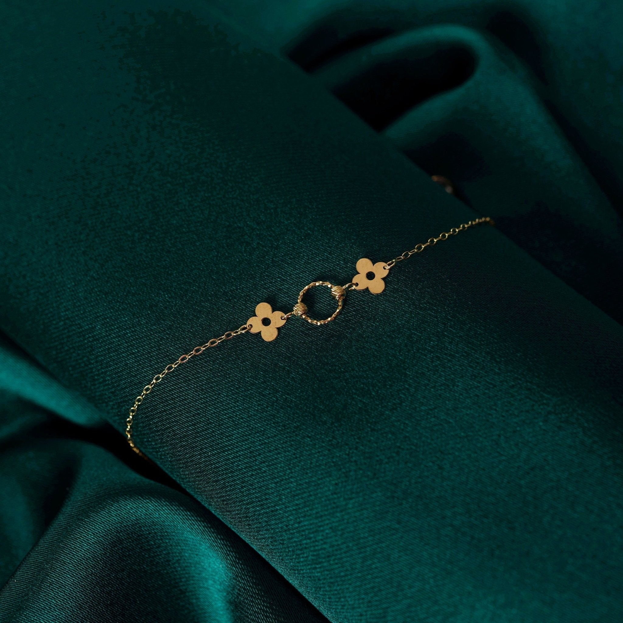 Fine gold-colored chain bracelet with two clover-shaped details and a textured circle, laid diagonally on dark green fabric.