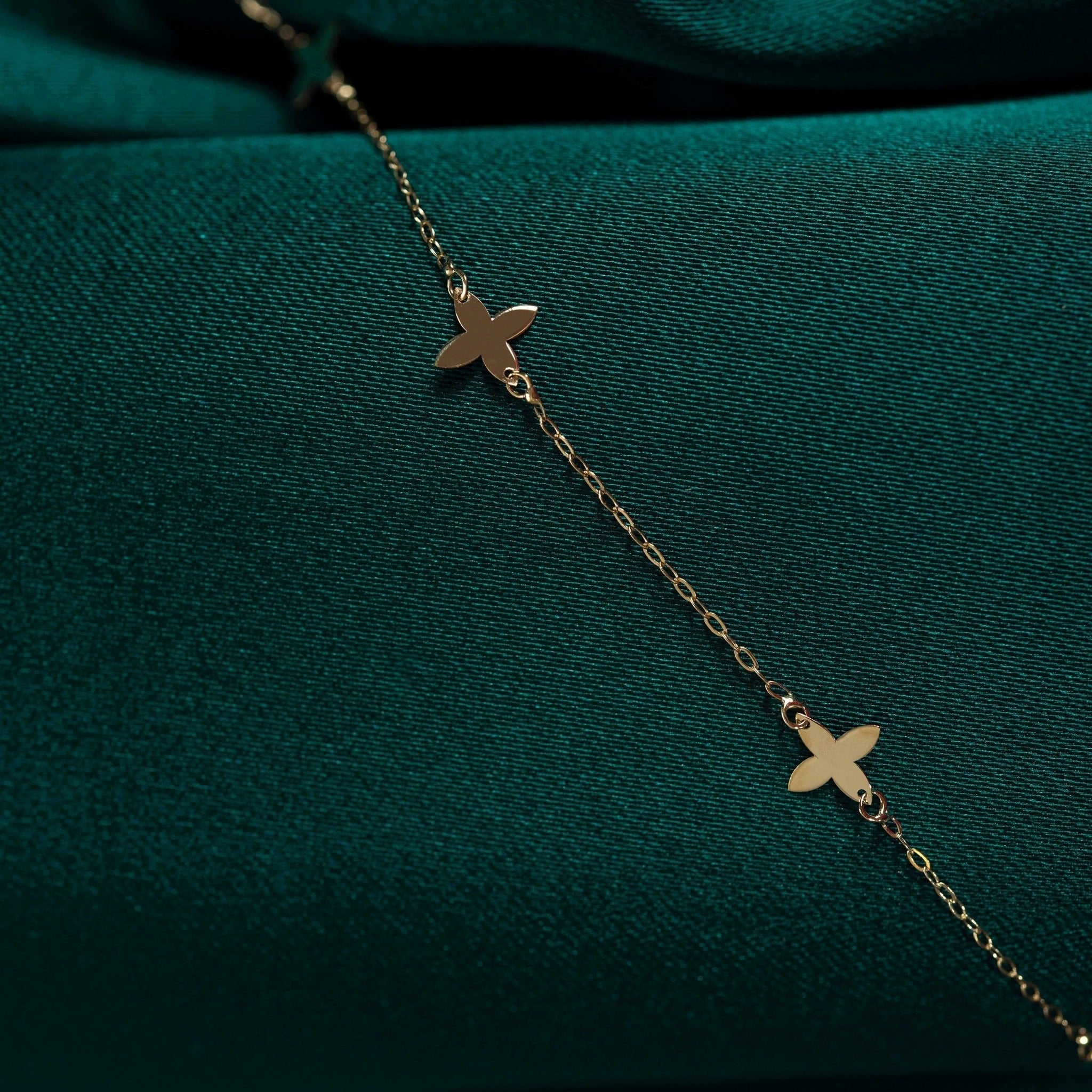 Close-up of a fine gold-toned chain bracelet with flat, four-petal clover shapes laid diagonally on dark teal fabric.
