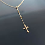 Gold-colored Y-shaped necklace with small beads and a cross pendant with rounded tips, resting on a dark surface.