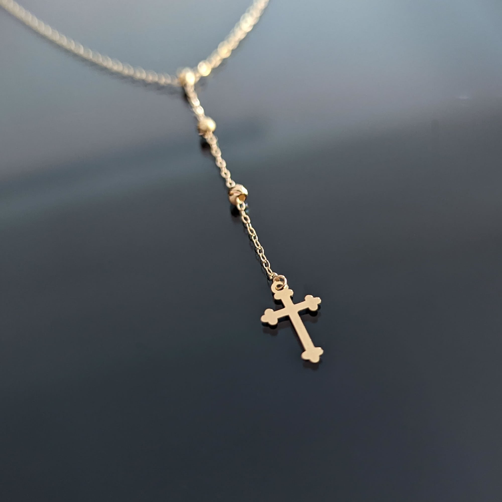 Gold-colored Y-shaped necklace with small beads and a cross pendant with rounded tips, resting on a dark surface.