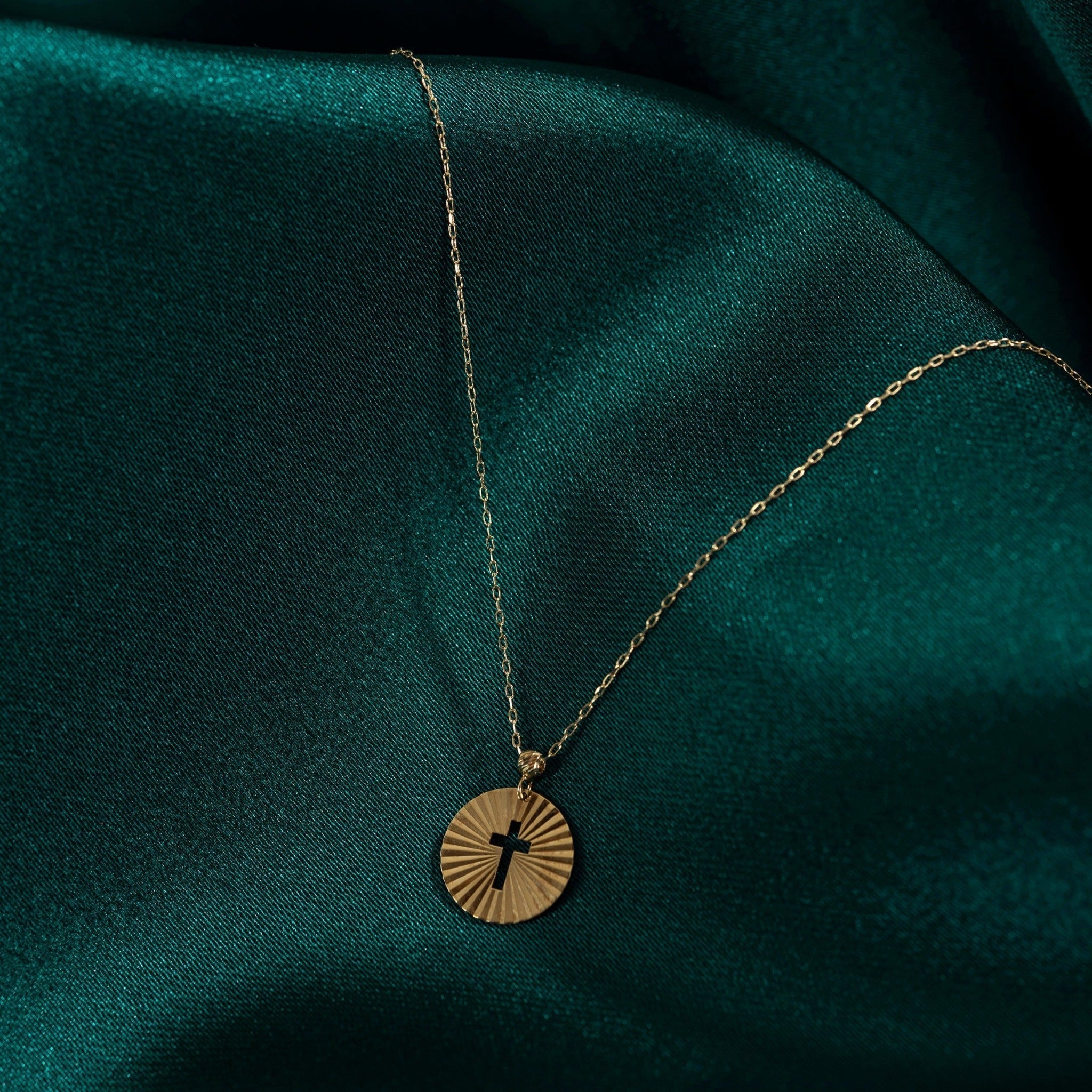 Gold-toned chain necklace with a round sunburst pendant and cross-shaped cutout, resting on dark teal fabric.