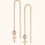 Pair of gold-tone threader earrings with long chains, clear faceted beads, and flat cross charms on a cream background.
