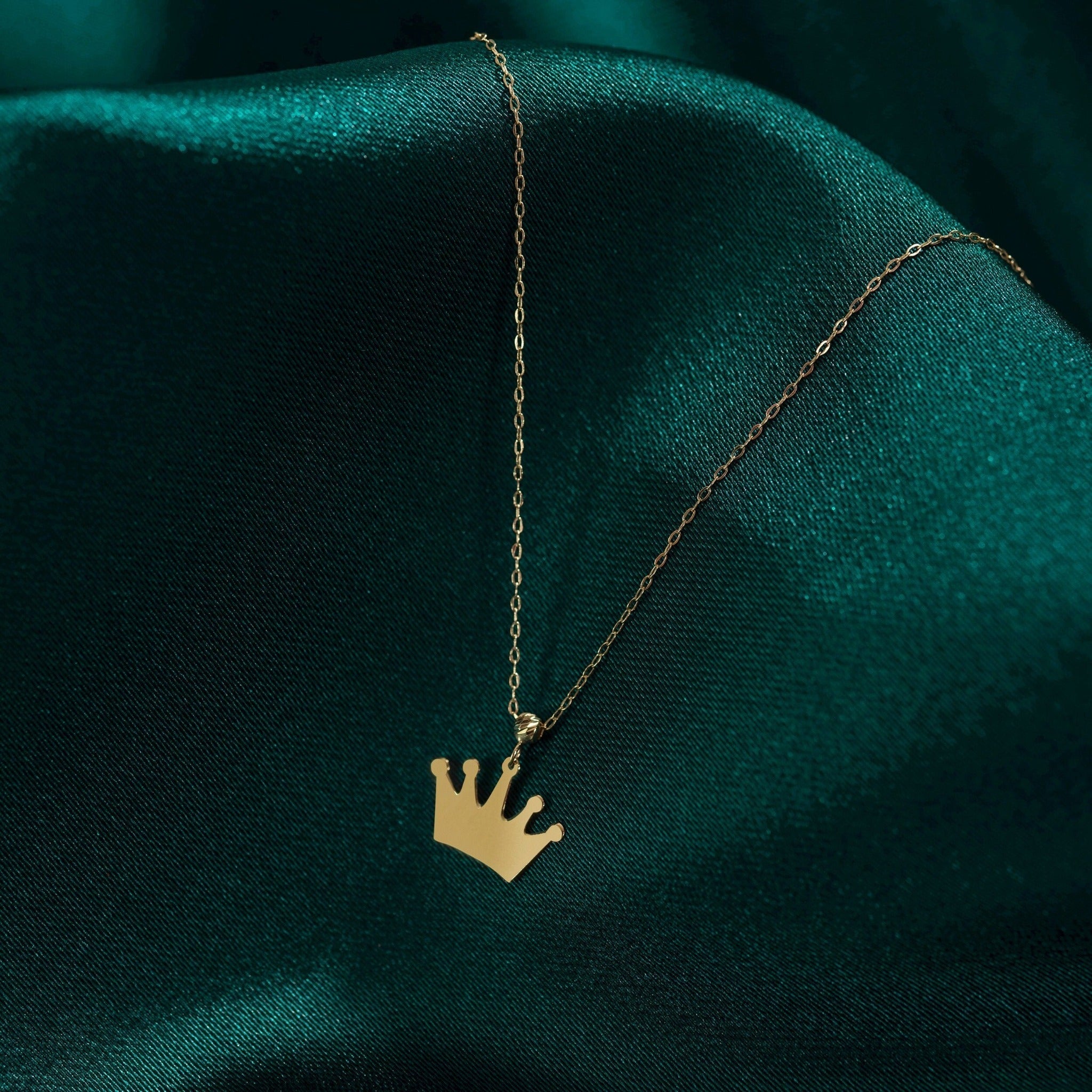 Gold-toned necklace with a flat, five-point crown pendant on a fine oval-link chain, resting on dark teal fabric.