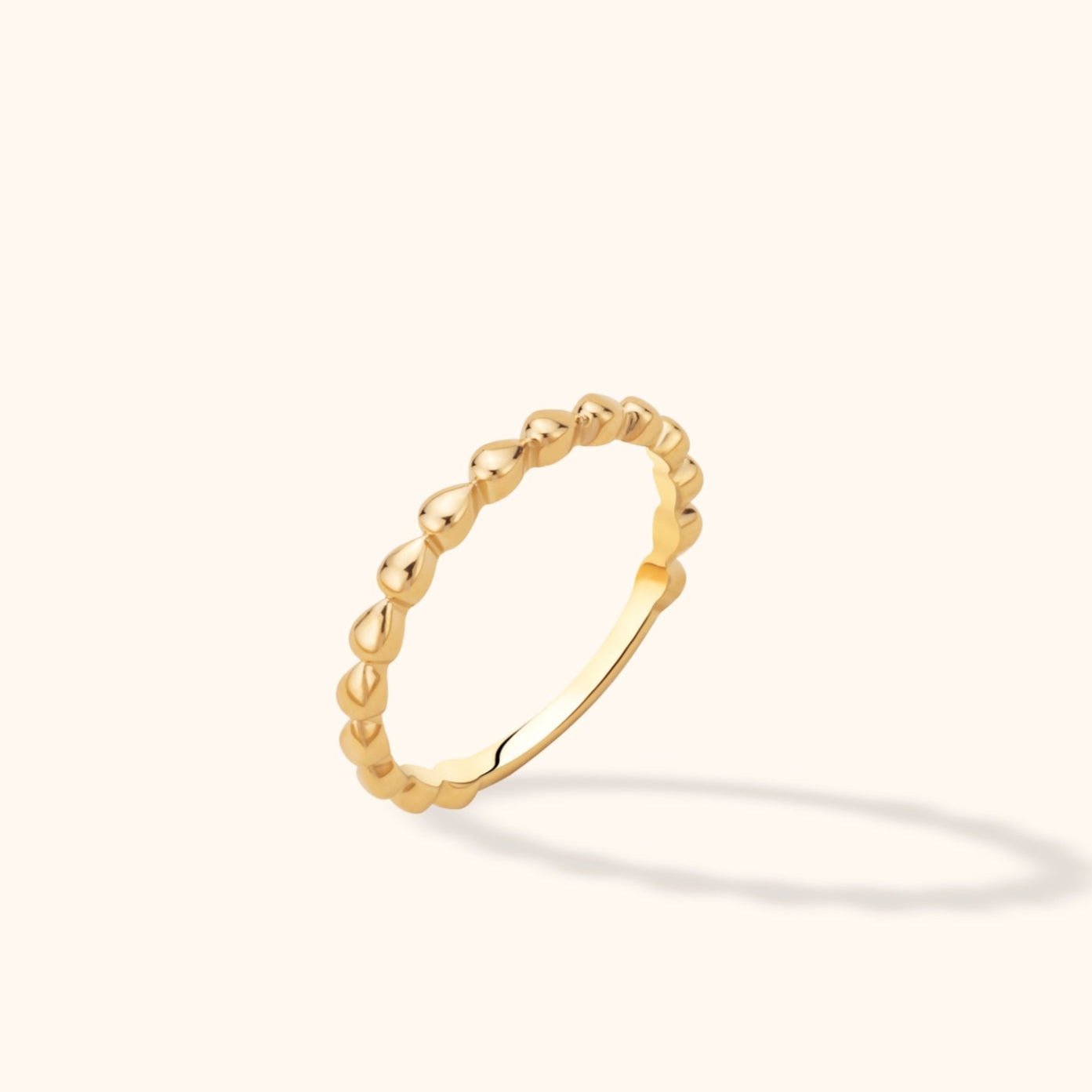 Slim gold-colored ring with repeating teardrop-shaped details, tilted upright on a cream background with a soft shadow.
