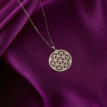 Gold-toned necklace with a round, openwork Flower of Life pendant and fine oval-link chain on glossy purple fabric.