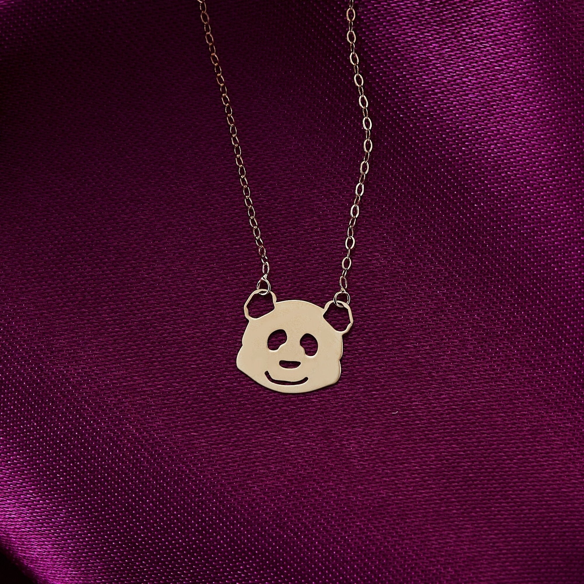 Gold-colored panda face pendant with cutout features on a fine oval-link chain, resting on purple fabric.
