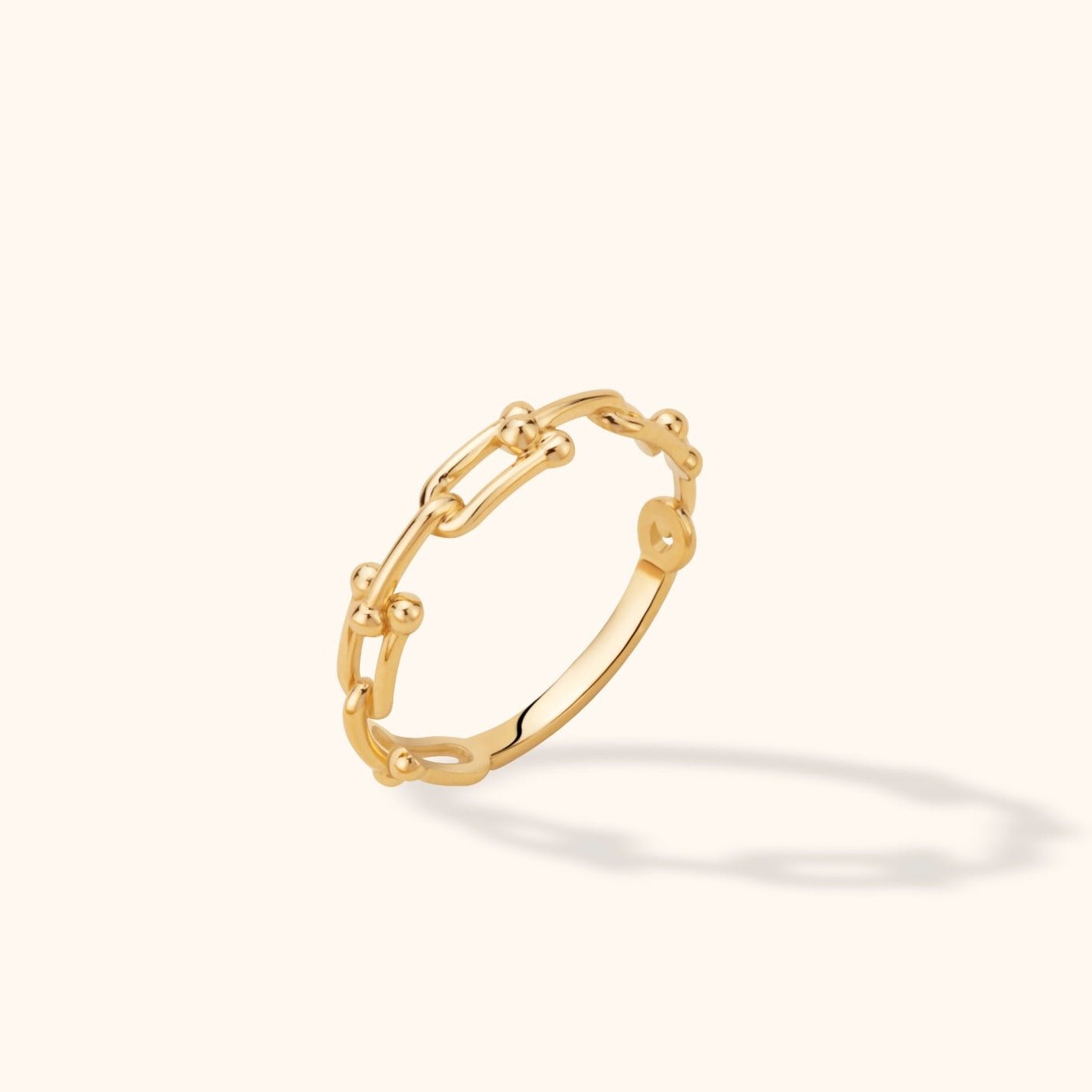 Gold-tone ring with elongated open links, small rounded accents and a smooth lower band, tilted against a cream background.