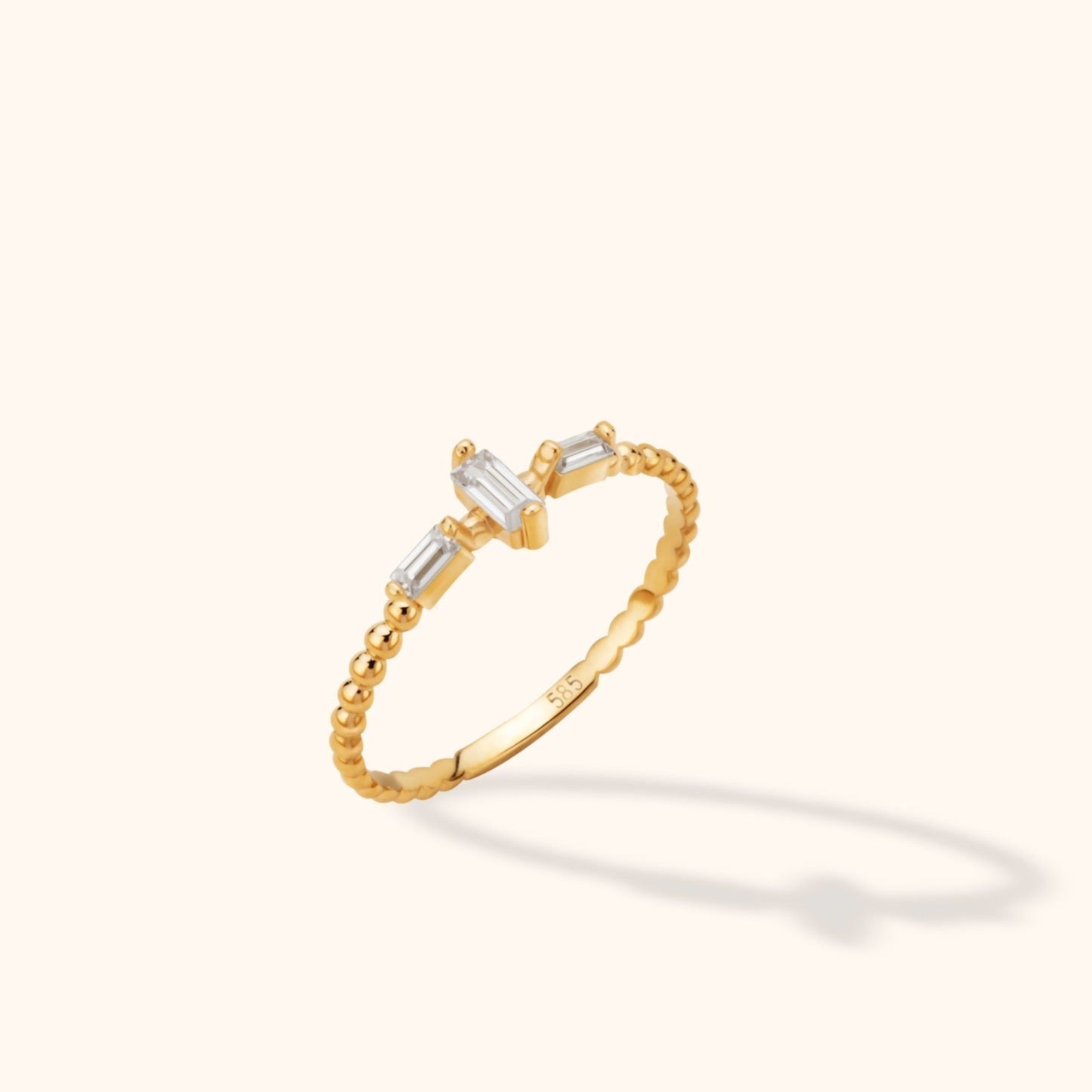Gold-colored ring with a slim beaded band and three clear rectangular stones, the center stone set crosswise, on a cream background.
