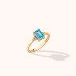 Gold-toned ring with a rectangular faceted blue center stone and clusters of small clear stones on either side.