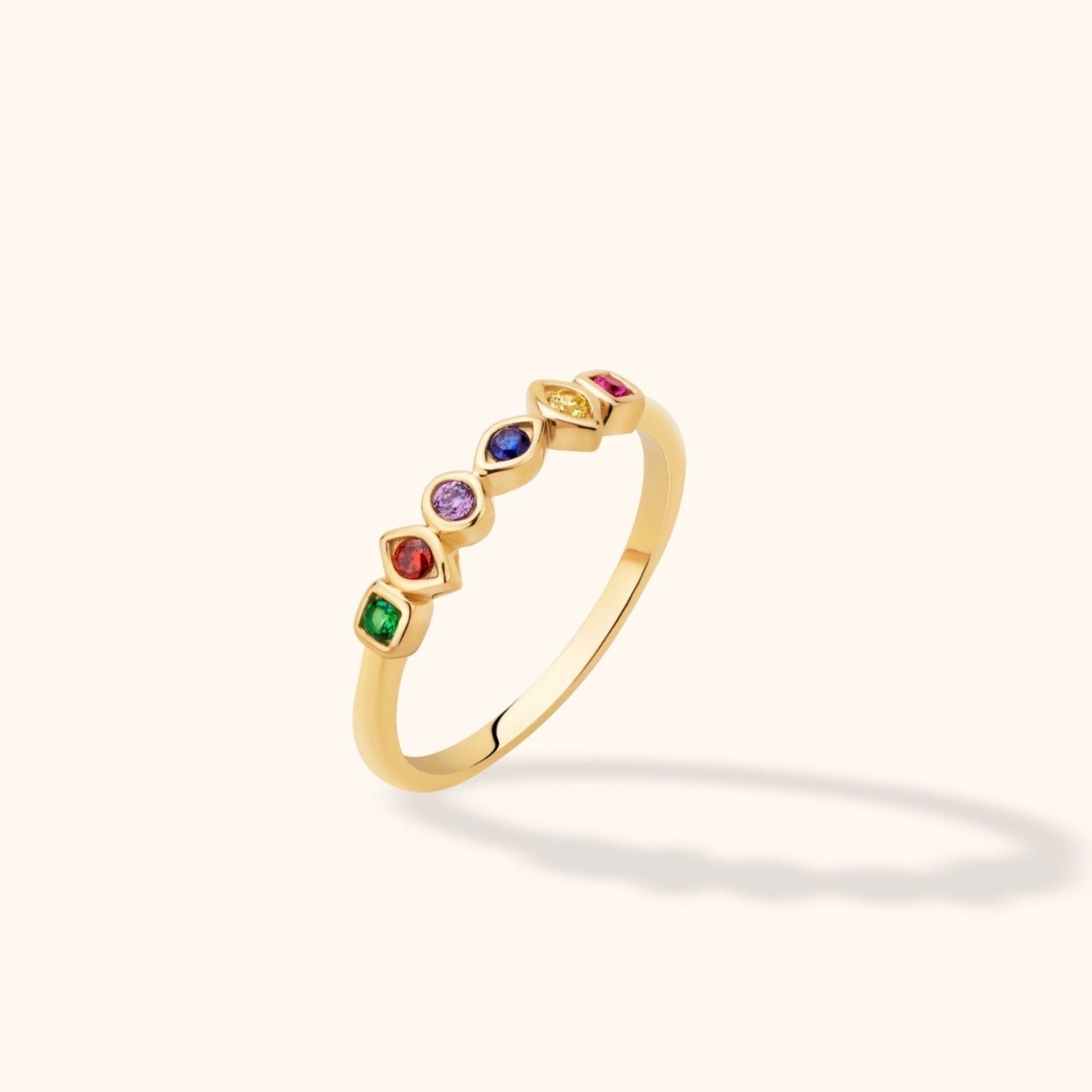 Slim gold-toned ring with six colorful accents in varied geometric settings, shown at an angle on a cream background.