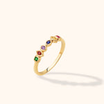Slim gold-toned ring with six colorful accents in varied geometric settings, shown at an angle on a cream background.