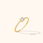 Slim gold-colored ring with a square clear stone in a smooth border setting, shown at an angle on a cream background.