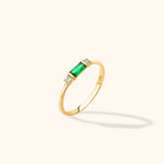 Slim gold-tone ring with a rectangular green center stone flanked by two round clear stones, shown at an angle on a cream background.