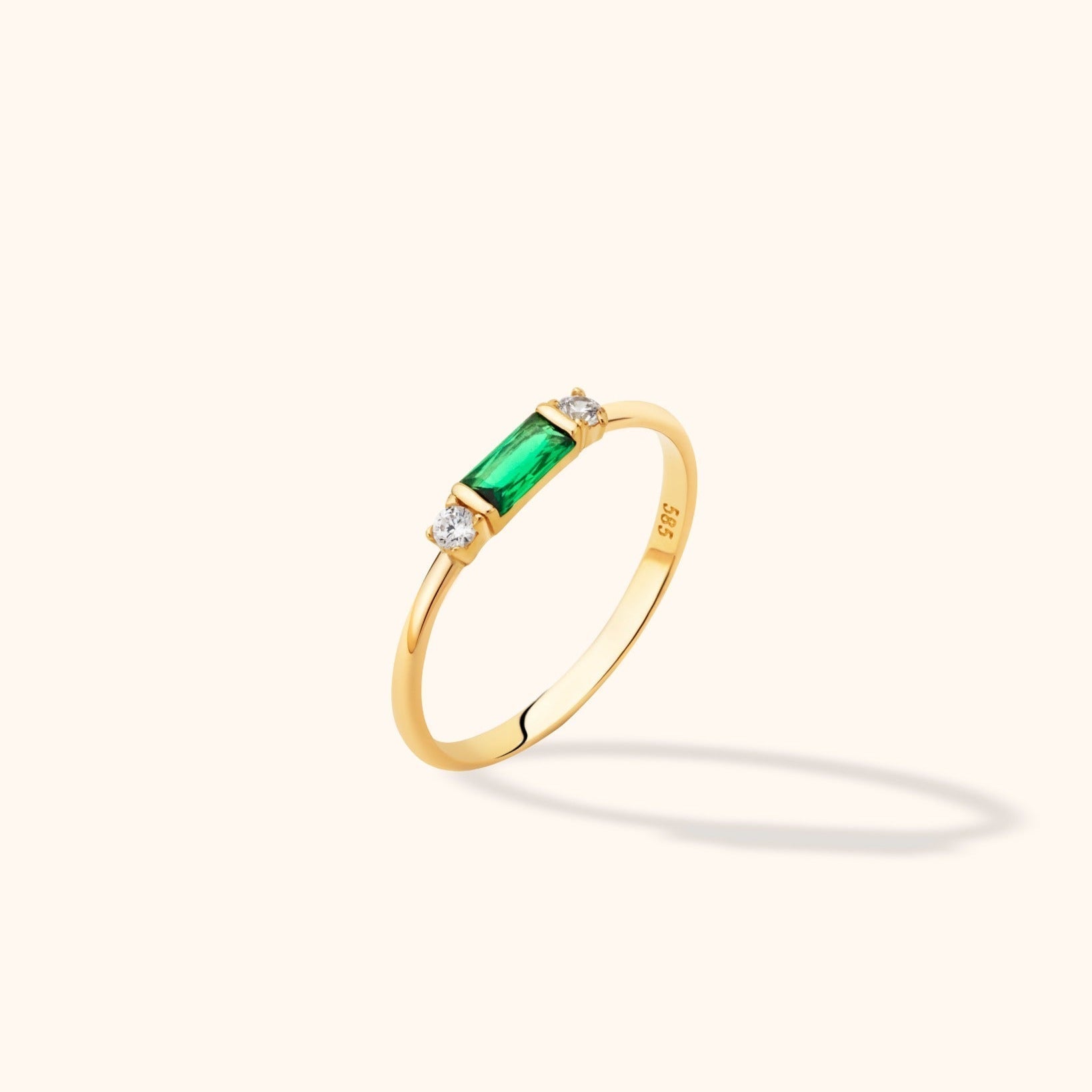 Slim gold-tone ring with a rectangular green center stone flanked by two round clear stones, shown at an angle on a cream background.