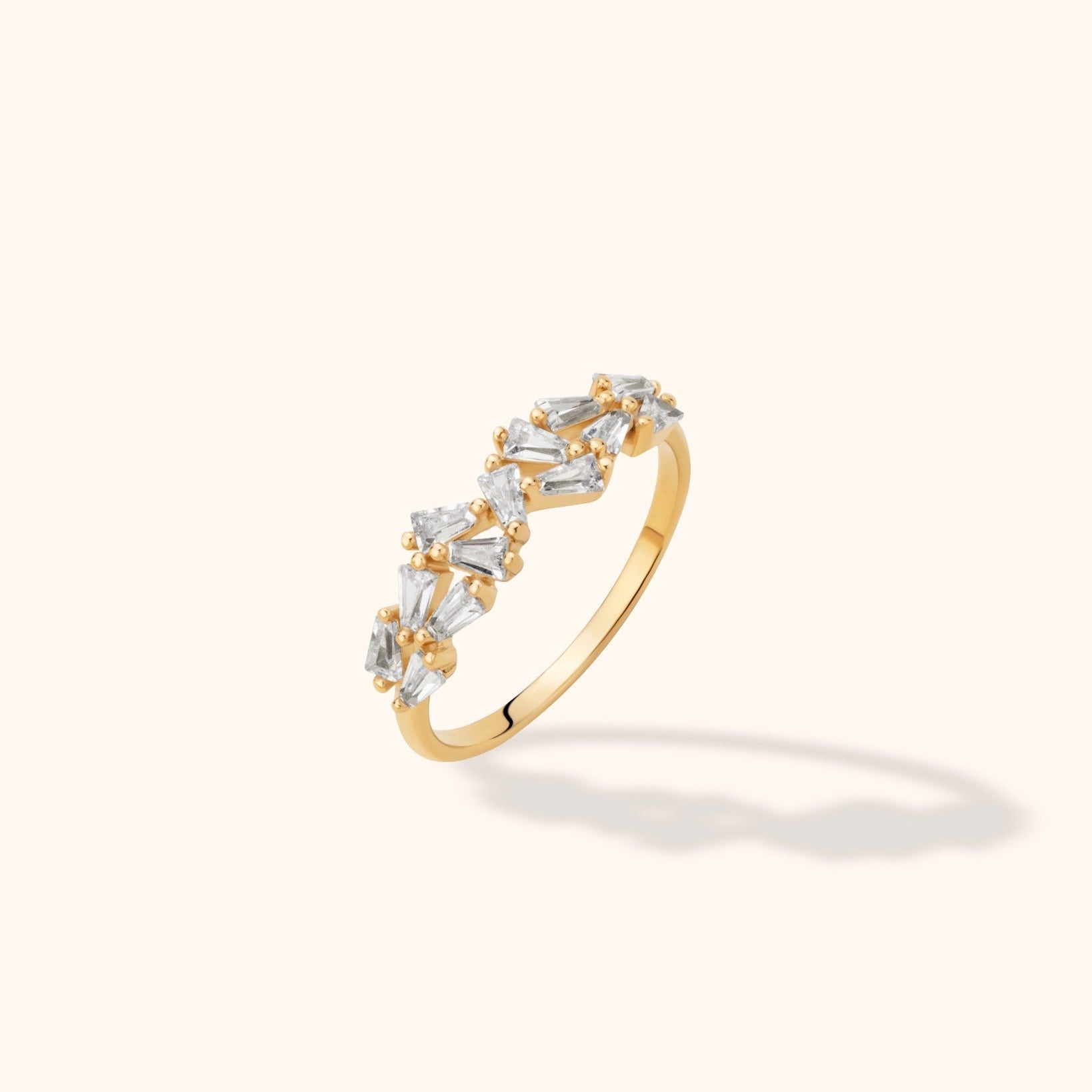 Gold-colored ring with clear, tapered rectangular stones arranged at varied angles across the top, shown on a cream background.