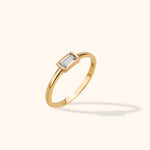 Slim gold-toned ring with a clear rectangular stone in a bezel setting, shown at an angle on a pale cream background.